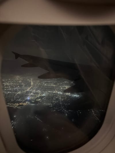 The image captures a view from an airplane window at night, looking down upon a sprawling city illuminated by countless lights. The wing of the aircraft is visible in the foreground, partially obscuring the cityscape. The city appears dense with buildings and a network of roads, crisscrossed by brighter lights that likely represent major thoroughfares. In the distance, the lights fade into a hazy glow, suggesting the vastness of the urban area. The overall impression is one of a vibrant, active city seen from a high vantage point. The image does not contain any information that would allow for the identification of the specific location within Köközek, Kazakhstan, or the presence of any individuals. The environment is consistent with an aerial view of a metropolitan area at night.