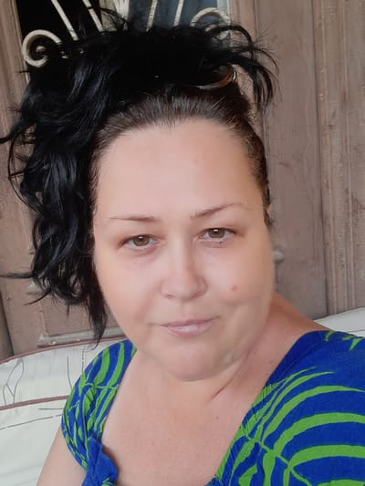 A medium, eye-level shot shows a woman with fair skin, light brown eyes, and dark brown/black hair. The hair appears tousled or wavy. She is wearing a top with a blue and green abstract pattern. The background includes what appears to be a brown wooden surface or wall, and the edge of a bed or upholstered piece of furniture is also visible. The setting or location in Cairo, Egypt is not evident from the provided photograph.