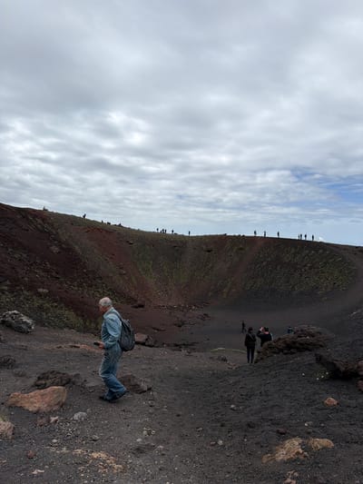 A landscape scene shows a volcanic crater near Nicolosi, Italy. The scene is set during the daytime under a cloudy sky. The foreground and lower right of the image are dominated by a dark, gravelly terrain with scattered rocks. An older man is walking on the terrain, wearing a light blue shirt, jeans, and a dark backpack. Several other people are walking along the edge of the crater in the background. The crater wall is steep and covered with dark soil and sparse vegetation.