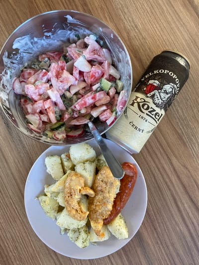 This is a top-down, eye-level, full-shot photograph of a meal placed on a wooden table. The meal consists of a large glass bowl filled with a salad of chopped tomatoes, cucumbers, and radishes mixed with a creamy white dressing, a small white plate with cooked potatoes, two pieces of breaded chicken, and a single sausage, and a can of Kozel beer. The can is positioned to the right of the salad bowl, leaning slightly. A metal fork is partially submerged in the salad. The overall lighting is bright and natural, suggesting it was taken indoors during the daytime. The wooden table has a visible grain pattern. There are no people in the image. The scene is set in the city of Swarzędz, Poland.