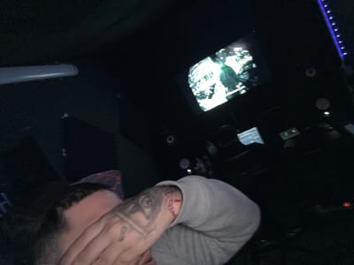 The scene depicts a dimly lit music recording or production studio, located in Kassel, Germany. In the immediate foreground, a person's upper body is visible, with their right arm bent and hand partially obscuring their face. This individual has short, dark hair and wears a light grey long-sleeved garment. Tattoos are present on their right forearm, featuring a large eye-like design, and a small figure-eight symbol on a finger. A red string bracelet is worn on the right wrist. In the midground, a second individual is seated at a desk, facing away from the camera, engaged with a computer monitor displaying what appears to be a digital audio workstation interface. Above the desk, on the back wall, a large screen displays a high-contrast, possibly monochromatic or green-tinted video featuring a human figure in a hooded garment. Studio monitors are positioned on either side of this screen. The room features dark soundproofing panels on the walls and is accented by a vertical strip of purple LED lighting on the right wall. A long, white linear light fixture is partially visible in the upper left.