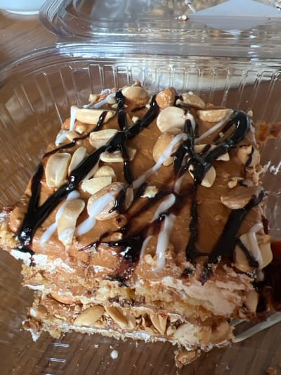 A close-up, overhead view depicts a multi-layered dessert slice housed within a clear, ridged plastic clamshell container. The dessert features a light brown top layer, possibly a frosting or ganache, heavily garnished with whole and halved peanuts. Dark brown and white sauces are drizzled in an irregular pattern across the top surface. The side of the slice reveals multiple horizontal layers, consisting of light-colored creamy strata interleaved with darker, crumbly layers that appear to contain crushed nuts or biscuit pieces. The plastic container rests on a light brown surface, likely wood, with a white object partially visible in the upper left background. The scene is located in Zeda Sakara, Georgia.