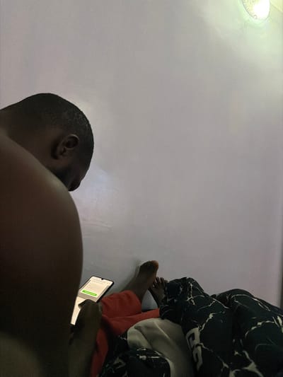 In Barakin Angundi, Nigeria, a dimly lit bedroom scene unfolds. A person, viewed from behind, is seated with their head bowed, engrossed in a smartphone held in their hands. The phone displays a webpage with fields for "Street Address," "City," and "Email," along with a green button labeled "Update Payment Method." Next to them, another person's legs are visible, draped in a dark-colored blanket with a white marble-like pattern. One foot is bare and rests on an orange surface, possibly a blanket or clothing. The background is a plain, light-colored wall with a subtle light fixture visible in the upper right corner, casting a soft glow. The overall atmosphere suggests a private, indoor setting, likely a home.