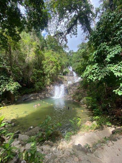 The image depicts a waterfall and pond in Tambon Thai Mueang, Thailand. The waterfall cascades down a rocky slope surrounded by lush green vegetation. A pool of water at the base of the waterfall is clear enough to reveal the pond’s floor. A yellow-buoyed rope stretches across the pool, possibly marking a safe swimming area. Two people are partially submerged in the water. A concrete or stone path edges one side of the pond. Trees and dense foliage dominate the surroundings.