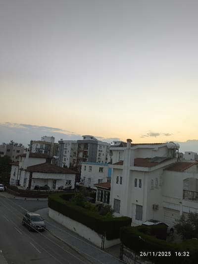 An elevated view captures a residential area in North Nicosia, Northern Cyprus / Cyprus, during late afternoon twilight on 26/11/2025 at 16:32. The sky transitions from a light gray overhead to a warm, pale yellow-orange at the horizon, with sparse, thin clouds visible in the mid-left.
In the immediate foreground, a multi-story, white stucco house with a reddish-brown tiled roof dominates the right side, featuring a prominent chimney, multiple windows (some with external shutters), a black metal fence, and a garage door. A light-colored vehicle is parked in its driveway, partially obscured by a hedge. Solar panels and a solar water heater are mounted on the roof.
To the left, a paved road with white lane markings curves slightly, flanked by a sidewalk and a well-maintained green hedge separating it from properties. A dark-colored hatchback car is situated on this road in the mid-ground. Further left and in the background, a light-colored car is partially visible parked on another street.
The middle ground and background consist of a mix of residential structures, including several multi-story apartment buildings with balconies, varying in design and color (some white, others with gray accents), interspersed with single-family homes with tiled roofs. Green trees and shrubbery are visible throughout the scene, primarily along property lines and between structures. No individuals are present.
