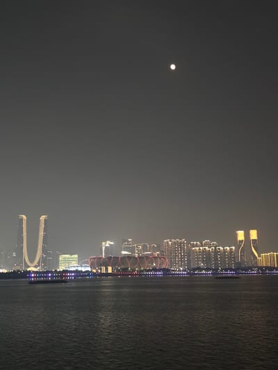 A night scene overlooks a wide body of water, likely a river or bay, with a city skyline in the distance. The sky is a hazy, dark gray, with a bright, circular moon positioned in the upper-middle section of the frame. The city lights are vibrant, outlining the buildings and creating a warm glow. Prominent structures include two distinct, curved skyscrapers on the left, illuminated with vertical lines of light, and a large, stadium-like building with red and white banding in the middle-ground. Further to the right, a cluster of tall buildings with uniform, illuminated windows stretches across the skyline. A long, low-lying promenade or embankment with a string of colorful lights runs along the waterfront, separating the water from the city. A barge or small boat is visible on the water, appearing dark against the slightly rippled surface. The overall impression is of a modern, illuminated cityscape at night. The location is Shangcheng District, China.