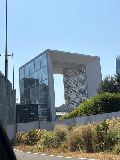 The image shows a view of the Grande Arche in Nanterre, France, on a clear, sunny day. The Grande Arche is a monumental arch-shaped building with a large open space in the center. It is characterized by its massive concrete structure and facade clad in granite and glass.
In the foreground, there is a section of a road, suggesting the photo was taken from a moving vehicle or a roadside. Adjacent to the road, there is a landscaped area with dry grasses and bushes, some of which are flowering with pale yellow blossoms. A grey concrete wall runs along the edge of the landscaped area, separating it from the buildings behind.
To the left of the Grande Arche, several other modern office buildings are visible. These buildings have glass facades that reflect the sky and surrounding structures. One of these buildings is taller and appears to be dark, possibly made of glass and metal. Another building to its right is lighter in color and has a more complex geometric design.
The Grande Arche itself is prominently featured in the center and right of the image. Its left side is a large, flat surface covered with reflective glass panes, mirroring the buildings to its left. The right side of the arch is also visible, revealing its internal structure with rows of windows. The massive opening of the arch is clearly defined by its concrete frame.
The sky is a uniform, light blue, indicating fair weather. There are no people visible in the image, and no specific actions or interactions are depicted. The scene is dominated by modern architecture and landscaping.