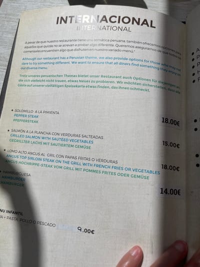 This is a close-up photograph of a restaurant menu, presumably taken by a customer. The menu is printed in black and white with a yellow accent. It is written in Spanish, English and German, and lists a selection of items from an international menu. The menu includes:
- "Solomillo a la Pimienta" (Pepper Steak) for 18.00 Euros
- "Salmon a la Plancha con Verduras Salteadas" (Grilled Salmon with Sauteed Vegetables) for 15.00 Euros
- "Lomo Alto Angus al Grill con Papas Fritas o Verduras" (Angus Top Sirloin Steak on the Grill with French Fries or Vegetables) for 18.00 Euros
- "Hamburguesa" (Hamburger) for 14.00 Euros
- "Menu Infantil" (Kids Menu) for 9.00 Euros.
The menu appears to be on a table with a hand visible in the bottom right-hand corner of the image. The image was taken in a restaurant in Las Palmas de Gran Canaria, Spain, at the coordinates 28.147, -15.43.