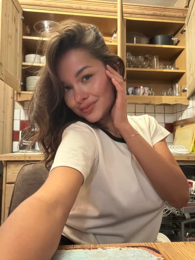 A young woman poses indoors with her hand near her ear. She has brown hair and a white t-shirt with a black collar. Kitchen cabinets with glassware, dishware, and cookware are visible behind her. A dishwasher can also be seen in the background. A portion of a tiled backsplash and a wooden countertop are also visible.