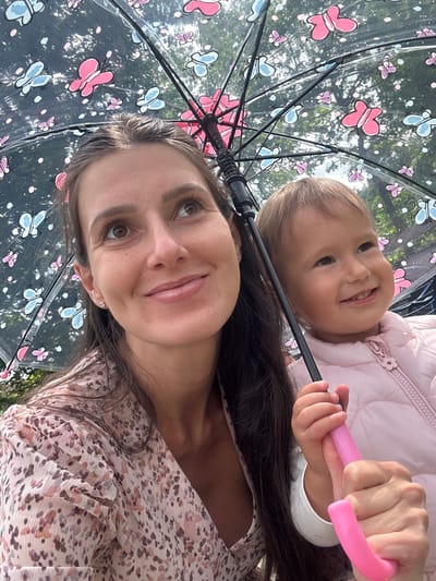 This close-up, low-angle shot captures a joyful moment between a woman and a young child, looking upwards through a clear umbrella. The setting is outdoors in Wisła, Poland, with bright green foliage and glimpses of a bright sky visible through the umbrella, indicating daytime.
In the foreground, a woman with long, dark hair and warm brown eyes looks slightly upwards and to the side, away from the camera, with a gentle, soft smile. She wears a light-colored, flowing top with a delicate floral or abstract pattern in muted pink, brown, and white tones.
Behind and above her, a young child with light brown hair smiles brightly, looking directly at the camera with an expression of pure happiness. The child is dressed in a light pink puffer jacket and holds the vibrant pink, curved handle of the umbrella with both hands.
The umbrella itself is a notable feature, made of clear plastic adorned with whimsical pink and light blue butterflies, along with scattered white dots. Its black frame and ribs are distinctly visible. The umbrella appears to be used playfully or for light protection, given the bright background.
The activity suggests a shared outdoor outing, possibly a walk or play, where the umbrella is a source of amusement. The emotions conveyed are warmth, affection, and joy, particularly from the child's beaming smile. There is no visible text in the image.