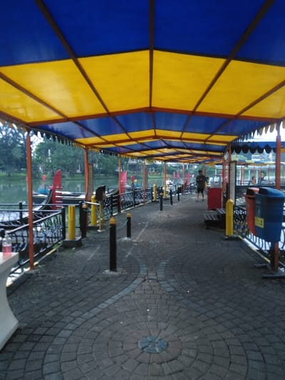 The image displays a covered walkway alongside a lake within La Sabana Park, Ciudad de San José, Costa Rica. Overhead, a long canopy features alternating blue and yellow rectangular fabric panels supported by a dark brown metal framework, extending into the background. The walkway is paved with dark gray interlocking pavers, incorporating decorative circular and curved patterns in lighter gray pavers. To the left, a black metal railing with yellow-capped bollards separates the walkway from the lake. Several red and dark-colored pedal boats are docked along the lake's edge. In the middle ground, a single male individual is visible walking away from the camera, wearing a dark shirt and light shorts. On the right side of the walkway, a wooden bench with a red frame, a blue trash receptacle with an orange lid, and a smaller red receptacle are present. Multiple black bollards with yellow caps are positioned along the walkway's edge. A white, low table or surface with a pink bottle on it is partially visible in the lower left foreground. The environment is outdoor, suggesting daylight conditions.