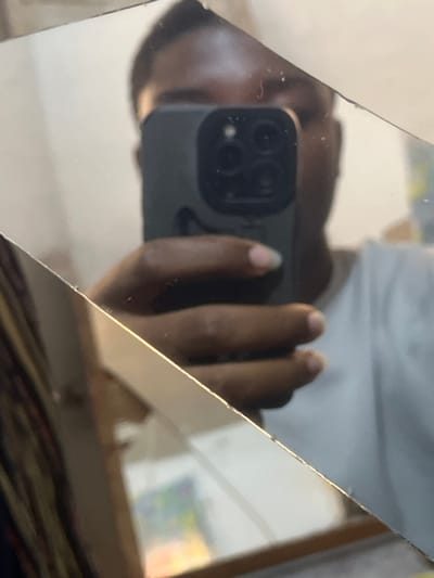 The scene depicts a reflection of an individual holding a dark-colored smartphone, possibly an iPhone due to its triple camera array and flash, in a fractured mirror or reflective surface. The reflection shows the upper portion of a person's face, primarily the forehead and eyebrows, with dark skin, obscured largely by the phone held in their right hand. The individual is wearing a white t-shirt. The right hand exhibits dark skin with natural-colored fingernails, some appearing discolored or soiled at the tips. The reflective surface is visibly broken by multiple diagonal cracks, segmenting the image. The background of the reflection appears to be an interior space with a light-colored wall and faint, multicolored elements, possibly artwork or decor, visible in the lower right quadrant. This event is observed in Bar Gada, Nigeria.