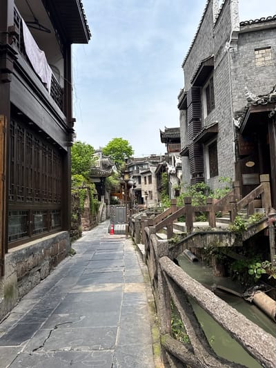 A narrow street runs through the city of Fenghuang, China. The buildings are traditional Chinese architecture, with grey brick walls and dark brown wooden accents. A canal runs alongside the street, separated by a wooden railing. A clothing rack can be seen hanging from a building. A utility cart is visible down the street.