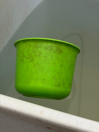 A bright green plastic bucket is floating in a bathtub filled with water. The bucket has a slightly worn appearance with some scuff marks and discoloration on its surface. The bathtub itself is made of a white material, and the water appears clear. The background is a muted, grayish-green color, possibly the wall of the bathroom. There are no people or other significant objects visible in the frame. The scene depicts a common household item in a domestic setting, likely a bathroom. This image is not associated with the city of Tupah, Indonesia.