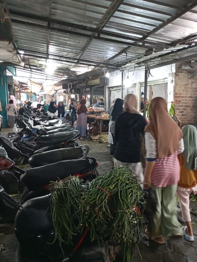 The scene is a bustling market in Kutawaringin, Indonesia, located at coordinates -6.953 latitude, 107.524 longitude. Multiple motorcycles are parked along the edge of the market. Green beans are piled on top of the motorcycles, with a few of the beans spilling onto the ground. The market itself is located in a partially covered open-air structure, with a metal roof and brick walls. There are several people walking through the market, some carrying bags, some holding items in their hands. One person wearing a light tan hijab, red and white striped shirt, and khaki pants is looking at the green beans while several other people stand in the background. Many of the people appear to be shopping and interacting with each other.