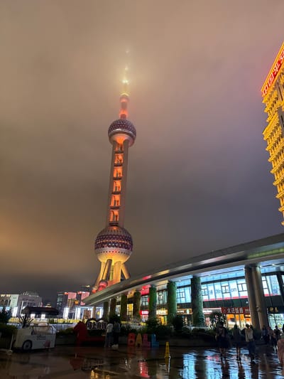 The image depicts a nighttime scene in Pudong, China, focusing on the Oriental Pearl Tower, an iconic landmark. The tower, illuminated with orange and yellow lights, pierces through a misty or foggy sky. The lower portion of the tower features large spherical structures decorated with lights, and a cylindrical section with illuminated rectangular openings.
In the foreground, a wide, wet plaza reflects the city lights. A modern building with a glass facade and vertical green accents runs along the right side of the frame, its windows displaying signs and advertisements. A prominent sign with Chinese characters is visible on this building. To the left of the Oriental Pearl Tower, a smaller building with illuminated windows and a light-colored facade is partially visible.
Several people are present in the scene, walking on the wet ground. An ice cream cart is parked on the left side of the plaza. In the middle ground, there are decorative elements including a heart-shaped sign with Chinese characters, a red sculpture, and large colorful letters spelling "BALL". The overall atmosphere is atmospheric and urban, characteristic of a city at night with some weather-related obscurity.