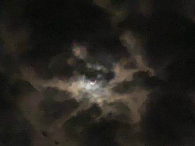 The image shows a crescent moon partially obscured by clouds in a night sky. The clouds are dark and appear to be wispy, with some illuminated areas where the moonlight shines through. The overall scene suggests nighttime, likely in a location with cloud cover. There are no people or discernible objects in the foreground. The image is a photograph of a celestial event, specifically a partial eclipse or a phase of the moon. The location context provided is Achara, Nigeria, which implies this celestial event was visible from that region. The mood is atmospheric and slightly mysterious due to the darkness and the way the clouds interact with the moonlight. No text is visible in the image.