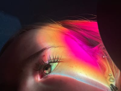 This is a close-up, low-angle shot of a person's eye and ear, illuminated by a vibrant spectrum of light that casts colorful streaks across their face and hair. The light creates a rainbow effect, with prominent bands of pink, orange, and blue. The person has multiple piercings in their ear, including a cluster of small, sparkling earrings near the top. Their hair, dyed a bright pink, is visible in the foreground, catching the colored light. The individual's eye is open, with long, dark eyelashes. The overall impression is artistic and surreal, with the play of light and color creating an otherworldly atmosphere. The scene appears to be indoors, with a dark background that emphasizes the illuminated subject. There is no visible text. The location context of Velykyi Sukhodil, Ukraine is not visually apparent from the image itself.