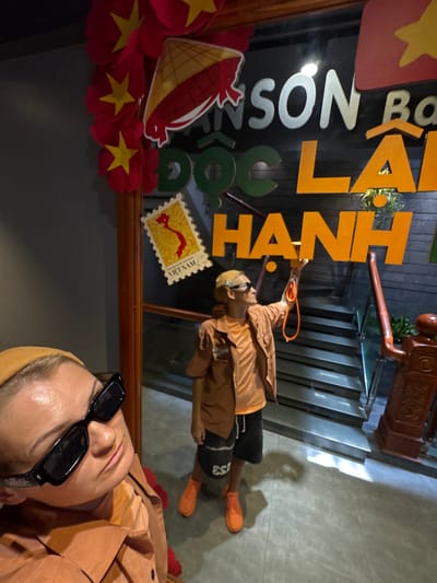 The image shows two individuals in front of a glass-fronted establishment. The person on the left, closer to the camera, is wearing a mustard-colored beanie and black sunglasses, with their face angled upwards and to the right. They are wearing an orange collared shirt. The person on the right is standing in front of a staircase, wearing a mustard-colored beanie, sunglasses, an orange short-sleeved shirt, and dark shorts. They are holding an orange cord in their right hand and reaching up towards a sign with orange lettering that reads "LA HANH". Green lettering above spells out "DOC". A portion of a larger sign with white lettering reads "SON BO". Decorative red and yellow elements are visible on the left side of the image, including a conical hat design. A yellow stamp with a red outline of Vietnam and the text "VIET NAM" is adhered to the glass. The flooring appears to be tiled. The environment suggests an indoor or semi-outdoor commercial space in Vietnam, possibly a restaurant or shop, located in South Quy Nhon Ward.
