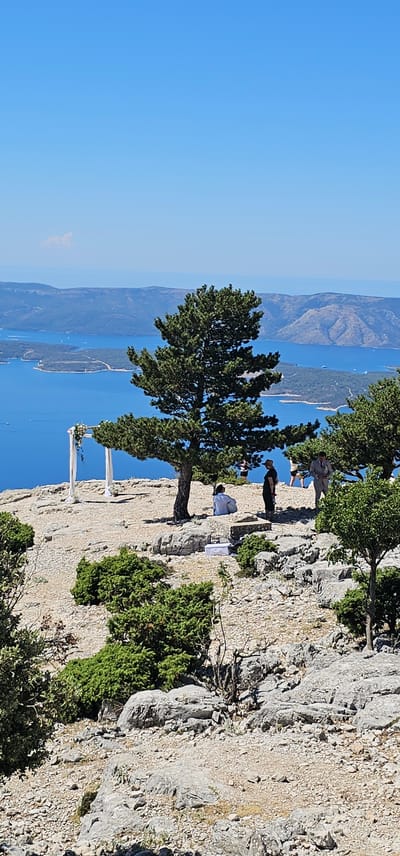 This is a vertical, eye-level, full shot of a wedding setup on a rocky clifftop overlooking a body of water and islands. The sky is clear and blue. In the foreground, there is a wedding arch draped with white fabric and adorned with flowers. A large evergreen tree stands to the right of the arch, with its branches extending over the rocky terrain. Several people are visible in the midground, some standing and some sitting. One person in a white shirt sits to the left of the tree, facing away from the camera. Another person in a dark outfit and a hat stands near the tree, facing towards the body of water. To the right of them, two more people are visible, one in a suit and one in a dress. A white box is placed on the ground near the seated individual. The clifftop is covered in rocks and sparse green vegetation. In the background, a vast expanse of blue water is dotted with small islands and boats. The overall scene suggests an outdoor wedding ceremony in a scenic, natural setting. The location appears to be the Vidova Gora peak on the island of Brač, in Općina Bol, Croatia, known for its panoramic views of the Adriatic Sea and surrounding islands.
