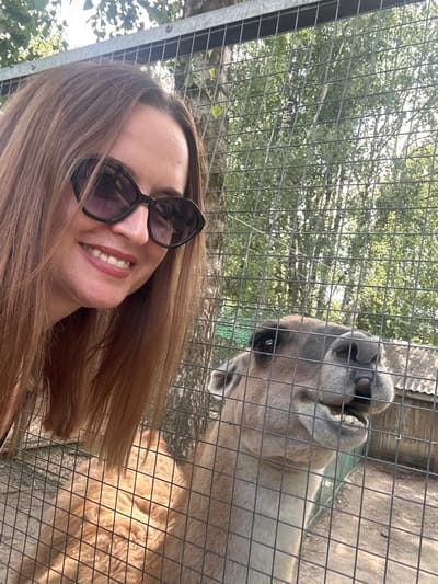 In a medium shot, a woman with long, auburn hair and sunglasses is smiling at the camera, leaning in towards a fence. Behind the fence, an animal, possibly a llama or alpaca, is looking into the camera with its mouth slightly open. The background consists of trees and a building, suggesting an outdoor enclosure, possibly at a zoo or sanctuary. The lighting suggests it is daytime, with sunlight filtering through the leaves of the trees. The woman's expression is happy and engaged, while the animal appears curious. The overall scene is casual and friendly, capturing a moment of interaction between a person and an animal. No visible text is present in the image. The location context is Medvedevo Urban Settlement, Russia.