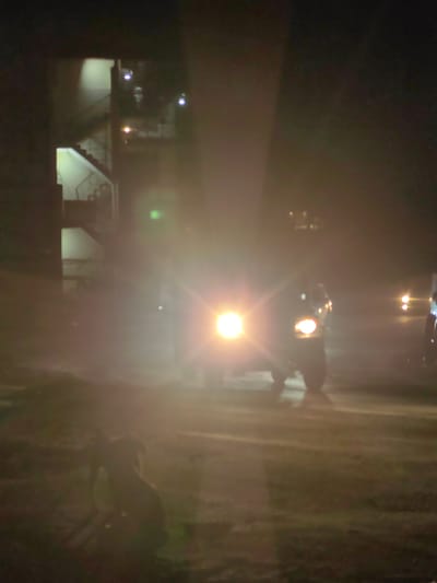 An outdoor, nighttime scene depicts an urban environment, possibly in Bahang, India, characterized by a building with external staircases illuminated by lights. In the foreground, a motorcycle with its headlight on is prominently featured, creating a strong lens flare that obscures details. To the left of the motorcycle, a dog is visible in silhouette. The overall lighting is dim and hazy, with multiple light sources contributing to a diffused and somewhat unclear image. The ground appears to be a dirt or gravel surface. The image captures a moment of low visibility, with the bright headlights of the motorcycle being the dominant light source.