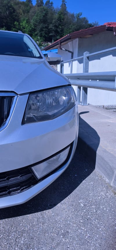 The image shows the front corner of a silver car parked outdoors. The car's headlight and part of the grille are visible. The car is parked on an asphalt surface, with a concrete curb on the right side of the frame. In the background, there is a white wall with a metal railing, and behind that, a red-tiled roof of a building and a hillside covered in green trees under a clear blue sky. The scene appears to be in a natural or semi-urban setting, possibly in Madan, Bulgaria, given the context. There are no people or animals in the image. The lighting suggests it is daytime, with strong sunlight casting a shadow from the car onto the ground.