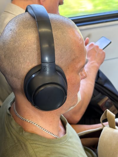 In a close-up, profile view of a bald man wearing black over-ear headphones and a green t-shirt, he is seated on public transportation, likely a train, as evidenced by the blurred green foliage visible through a window in the background. The man is holding a black smartphone in his right hand, interacting with it. A silver chain necklace is visible around his neck, and a white medical mask is partially covering his mouth and chin. The scene appears to be during daylight. The background suggests an urban or suburban environment with a hint of green. The image does not provide any specific indicators to pinpoint a location within the city of Shcherbinka, Russia.