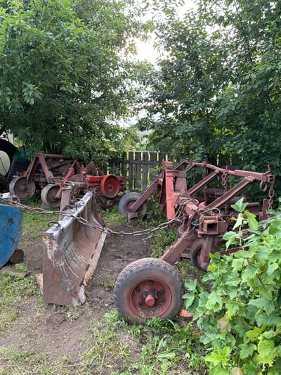 This is a tilted, medium-wide shot of old, rusty farm equipment, possibly a plow or cultivator, and a bucket attachment, set against a backdrop of lush green trees and foliage. The equipment is made of metal, painted red and brown, and appears weathered and worn, with visible rust and chains. The bucket attachment is large and scoop-like, also showing signs of age and use.
The scene is outdoors, in what appears to be a rural or garden setting. The ground is a mix of dirt and grass, with some weeds growing around the equipment. The trees are dense and leafy, suggesting a warm season, possibly summer. Sunlight filters through the leaves in some areas, indicating it is daytime. The weather appears overcast or cloudy, with diffused light.
There is a rustic wooden fence visible in the background, adding to the rural atmosphere. The overall impression is one of disuse and abandonment, with the old machinery left to the elements. There are no people visible in the image. The setting is consistent with a rural yard or farm in the region of Chaykovsky, Russia, suggested by the lush greenery and traditional fencing. There is no visible text in the image.