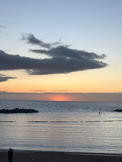 The image shows a sunset scene at the coast. A large, dark cloud formation is visible on the left side of the image, contrasting with the lighter, orange-hued sky. The sun is setting on the horizon, creating a visible line where the sky transitions to the ocean. The water appears calm with ripples, and the light is reflecting in the water, creating a shimmering effect. Along the shoreline, a breakwater with rocks is visible. On the lower right corner, a person is visible on the beach, seemingly observing or photographing the sunset. The location is likely near the coast of Mogán, Spain, given the presence of the ocean, breakwater, and the characteristic sunset colors.