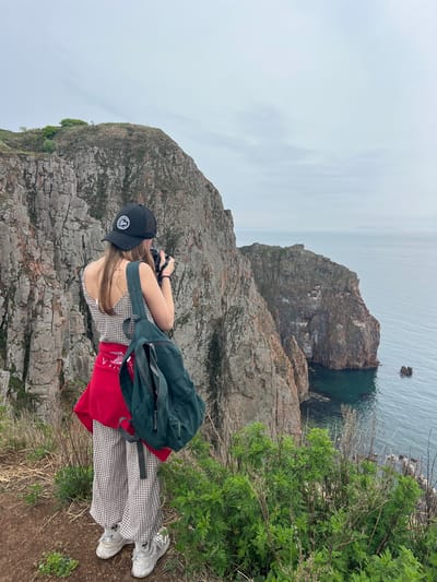 A woman stands on a cliff overlooking the sea in Russkiy, Russia. She is wearing a black baseball cap, a checkered top, a red sweatshirt tied around her waist, checkered pants, and white sneakers. She has a green backpack on. She is holding a camera to her eyes, taking a photograph of the scene. The cliff is rocky and barren, with some greenery growing on the edge. The sea is calm and gray. The sky is overcast.