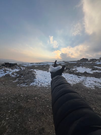An arm, clad in a black padded jacket and a black glove, extends from the lower right of the frame, holding an irregular clump of white snow. The scene depicts a desolate, open landscape in Talfita, Syria, under a partly cloudy sky with the sun visible through clouds in the upper right, casting a warm glow. The ground is largely covered in dark, rocky soil and gravel, with scattered patches of white snow. In the midground, to the left, are multiple piles of light-colored rubble, consistent with demolition debris. Adjacent to this debris, a rusted, dark-colored vehicle, possibly a truck, is partially visible. Further into the midground and background, the terrain continues with similar rocky and snowy patches. On the distant left horizon, faint structures resembling power line towers are visible. To the far right, on a slight rise, several multi-story structures, possibly residential buildings, are observed. No other individuals are present.