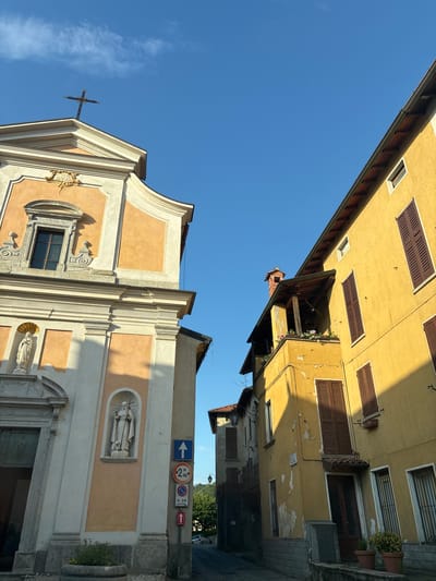 The image captures a street scene in Orino, Italy, on a clear, sunny day with a bright blue sky. On the left, the facade of a peach-colored church stands prominently, adorned with a cross at its apex and religious statues. The church features architectural details like ornate window frames and a large arched doorway. To the right, a vibrant yellow building with brown shutters lines the street. Its weathered exterior shows signs of age, with peeling paint in places. A small balcony with potted flowers overlooks the street, and a brick chimney protrudes from the roof. The street itself is narrow and paved, with a few traffic signs visible, including one indicating a height restriction. Potted plants are placed along the base of the yellow building. The lighting suggests it is either early morning or late afternoon, with long shadows cast across the scene.