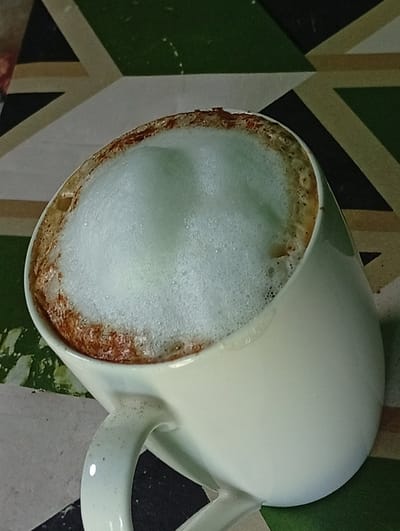 A close-up shot shows a white mug filled with a foamy beverage, likely coffee or a latte. The foam is light and airy, with a slightly off-white, pale green hue. Around the rim of the mug, there's a thin layer of darker brown, indicating where the liquid meets the foam, possibly from coffee residue. The mug itself is plain white with a speckled pattern, featuring a curved handle.

The mug is placed on a surface with a geometric pattern. This surface consists of alternating colored sections: dark green, light beige, and dark gray or black. These sections form diagonal stripes and triangular shapes, creating a dynamic and visually interesting background. The overall lighting suggests an indoor setting, possibly during the daytime, as there are no strong shadows.

The image is cropped to focus primarily on the mug and its contents, with the background providing context without being overly distracting. There is no visible text in the image. The setting appears to be a table or countertop in a home or cafe. The image is static and does not depict any ongoing activity or event beyond the preparation and presentation of the beverage. The composition and lighting are simple, highlighting the texture of the foam and the design of the surface.
