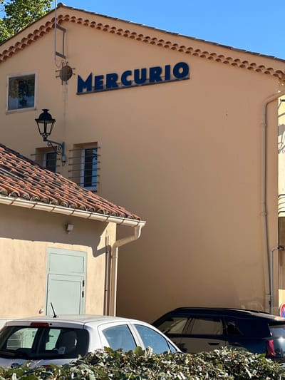 The image captures a building with a beige facade and a tiled roof, featuring the blue lettering "MERCURIO" on its upper section. Below the sign, there is a window with bars and a street lamp. To the left, another section of the building has a tiled roof, a window with bars, and a light blue door. In the foreground, the tops of cars are visible, including a white car with a rear spoiler and an antenna, and a dark-colored SUV. Lush green foliage partially obscures the lower part of the image. The scene is set against a clear blue sky. The architecture and the name "MERCURIO" suggest a location in Saint-Tropez, France.
