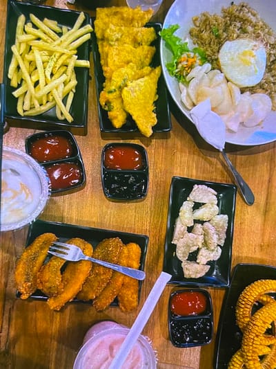The image captures a top-down view of a wooden table laden with various Indonesian dishes, likely in Cadek, Indonesia. In the upper left, a black rectangular dish holds a pile of golden french fries. To its right, another black dish contains several pieces of fried tempeh, a popular Indonesian soy product. Dominating the upper right quadrant is a white oval plate filled with fried rice, topped with a fried egg and a small garnish of shredded vegetables and lettuce. Accompanying the fried rice are several translucent, disc-shaped crackers.

Below the french fries, a small black dish is divided into two compartments, one filled with red sauce (likely ketchup) and the other with a dark sauce (possibly chili or soy sauce). To the left of this is a clear plastic container with a white, creamy substance, possibly a dip or yogurt.

In the lower left, a black rectangular dish displays several pieces of fried food, appearing to be banana fritters, with a silver fork resting on top. Adjacent to this, a partially visible pink drink with a white straw is present. Below the fried rice plate, another black dish holds several pieces of pale, fried snacks. Finally, in the lower right corner, a black rectangular dish features a portion of corn on the cob, sliced into rings. The overall scene suggests a casual meal setting with a variety of fried and savory items.