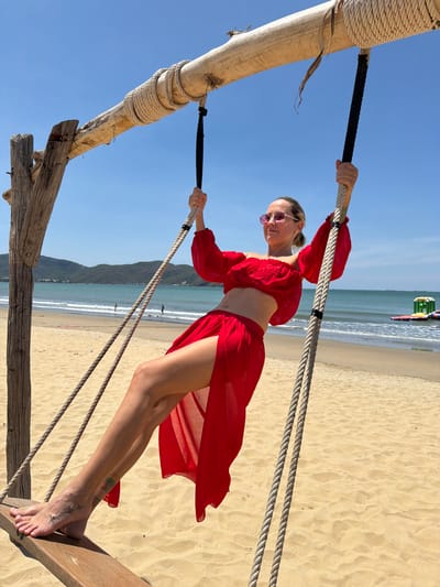 A woman in a red two-piece outfit and sunglasses is on a wooden swing on a sandy beach with the ocean and hills in the background. The swing is suspended from a wooden beam by ropes. The woman is holding onto the ropes with her hands and her legs are extended. She has tattoos on her feet. In the distance, there are two people walking on the beach and some floating structures in the water. The sky is clear and blue. The location appears to be a beach in Quy Nhon Ward, Vietnam.
