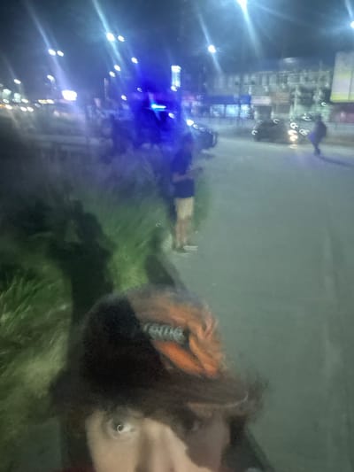 A blurry, rotated image shows a person lying on grass near a city at night. Bright blue lights illuminate buildings across the frame. The person is wearing dark shorts, a black shirt, and an orange jacket. Raindrops or water droplets are visible on the lens, contributing to the blurry effect. The image is distorted, making details difficult to discern.