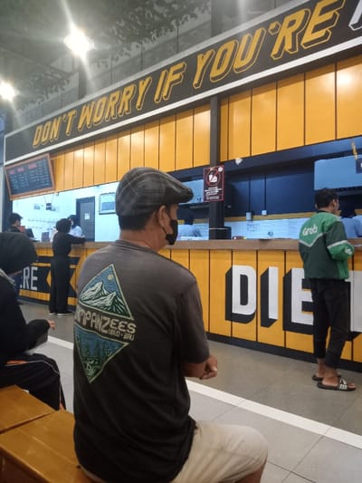 This image depicts an indoor dining or waiting area within a casual eatery in Nganjuk, Indonesia. The primary subject is a male figure in the foreground, seated on a wooden bench with his back partially to the camera, looking towards a service counter. He wears a grey plaid flat cap, a black face mask, a dark grey t-shirt featuring a mountain and forest graphic with "CAMPANZEES UBUD - BALI" text, and light-colored shorts. His hands are clasped in front of him. To his immediate left, another person in dark attire is partially visible, also seated on the bench.
The background features a bright yellow and black-paneled service counter area. Above the counter, a large black sign with yellow 3D lettering reads "DON'T WORRY IF YOU'RE". A menu board displaying white text on a dark background is visible to the left. A black sign with red and white elements, likely a warning or informational notice, hangs centrally above the counter. Behind the counter, several individuals, presumably staff, are present in a well-lit food preparation or service area. To the right, another male figure, a delivery driver, stands facing the counter. He wears a green "Grab" uniform jacket, dark cropped pants, and black and white striped slide sandals. The floor is light-colored tile with a white line running parallel to the counter. Overhead, bright lights illuminate the space, and the ceiling features decorative elements resembling artificial foliage. The counter facade on the lower right displays partial white 3D block letters spelling "DIE".