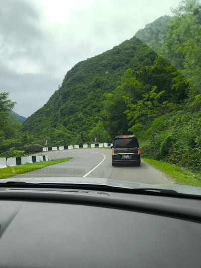 This image depicts a scenic view from inside a vehicle, likely traveling on a mountain road in the vicinity of Gamoghma Nogha, Georgia. The road curves to the left, bordered by a black and white concrete barrier on the left and lush green vegetation on the right. A dark-colored minivan is visible ahead on the road, with its brake lights illuminated. The landscape is dominated by a steep, densely forested mountainside to the right, partially obscured by clouds or mist at higher elevations. The sky is overcast with gray clouds, suggesting a cool or rainy day. The foreground shows the dashboard of the vehicle from which the photo is taken. Road signs indicating a sharp curve are visible along the barrier. The overall impression is one of a journey through a verdant, mountainous region.