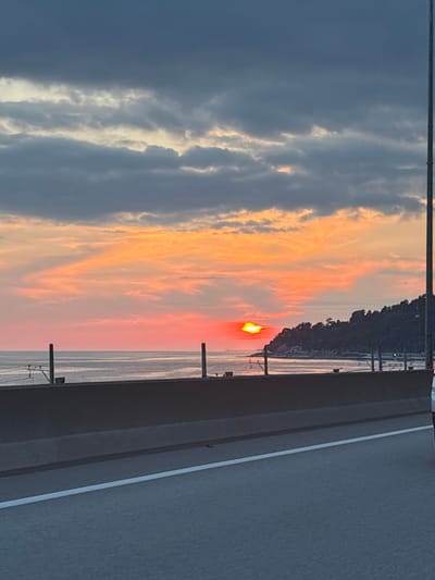 The image depicts a sunset over a body of water, likely a sea or large lake, with a mountainous coastline in the distance. The sky is a dramatic display of color, with vibrant orange and red hues from the setting sun bleeding into softer pinks and yellows, contrasted by darker, moody clouds.

In the foreground, a paved surface with a white line suggests a road or walkway, possibly a bridge or elevated path, offering a vantage point over the scene. The surface appears to be grey asphalt. To the left of this paved area, a metal railing or barrier is visible, hinting at safety measures for those overlooking the water. Two vertical posts, also possibly part of a railing or support structure, stand in the water.

The time of day is clearly sunset, indicated by the low angle of the sun and the intense colors in the sky. The weather appears to be clear enough for a sunset, though the presence of clouds adds depth and drama to the sky. The location cues, such as the coastline and the water, point towards a coastal or lakeside setting in Adlerskiy, Russia. There is no visible text in the image. The overall mood is serene and awe-inspiring, capturing the natural beauty of a sunset. There are no people or specific objects that dominate the scene, with the focus being on the landscape and the celestial event.