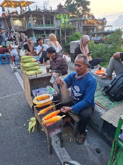The scene is a street food vendor in Kecamatan Piyungan, Indonesia (latitude: -7.846, longitude: 110.48). A man is grilling corn on a charcoal grill, while another man sits on a wooden stool to his right. The man is using tongs to turn the corn, which is on skewers. There are several other people in the background, including two women wearing headscarves, and a young man wearing a white shirt and black pants. There are bags of corn stacked on a wooden table beside the grill. The scene is set on a paved road with a concrete curb. The background shows several buildings and trees.