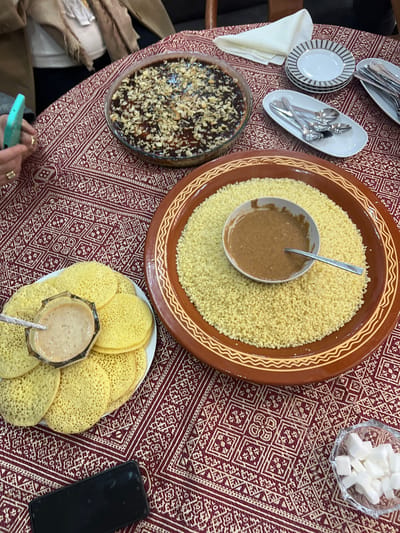 The image presents an overhead view of a table adorned with a vibrant red and gold patterned tablecloth, showcasing a spread of traditional dishes, likely from Ait Ouallal, Morocco.
At the center, a large, ornate terracotta-colored serving dish holds a substantial quantity of fine, pale yellow grains, resembling couscous. A smaller white bowl, containing a thick, brown sauce or dressing with a spoon, is nestled in its middle. To the left, a clear glass baking dish features a dark, glistening dessert generously topped with chopped nuts, possibly a nut cake or pastry. Nearby, a stack of small, striped plates and an elongated white dish filled with an assortment of serving spoons and forks are laid out. A folded white napkin completes this section of the table.
On the lower right, a plate is laden with several light yellow, round pancakes, characterized by a porous, spongy texture, commonly known as Baghrir in Moroccan cuisine. A small, faceted glass bowl, holding a light brown liquid – possibly honey, syrup, or Amlou – along with a spoon, rests on top of these pancakes. In the upper right corner, a clear glass bowl is filled with white sugar cubes.
The hands of at least one individual are visible on the left, holding a light blue smartphone, while a black smartphone is visible on the right, indicating a casual setting. The intricate geometric patterns of the tablecloth, coupled with the distinctive food items and serving ware, strongly suggest a Moroccan cultural context. The arrangement implies a shared meal or gathering.