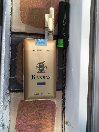A pack of "Kansas" brand cigarettes, with "American Blend" text and a coat of arms graphic, rests on a tiled surface. The pack is gold colored and stands upright. To the left of the pack are two cigarette filters, partially obscured. To the right of the pack is a black and green lighter with the "FOX" logo. The tiled surface appears to be outdoors, with a white vertical structure to the right. The scene is set in the city of Rivne, Ukraine.