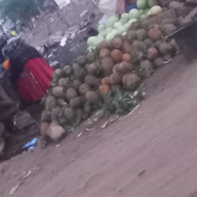 The image appears to be taken from an elevated perspective in Barnawa, Nigeria. A vendor with a red head covering is shown near a display of produce. The produce includes pineapples, some appearing green and unripe, others with a more mature brown coloration, and a separate collection of green, spherical fruits, potentially limes. The produce is arranged near what appears to be a concrete or stone surface. Other items and debris are visible in the lower left quadrant.