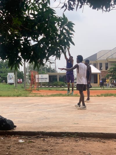 In Ukukwa, Nigeria, on an outdoor basketball court, three young men are engaged in a game. One player in a purple jersey and shorts jumps to shoot the ball, while another player in a white sleeveless shirt and black shorts stands nearby, seemingly observing the play or preparing to defend. A third player, wearing a black tank top and shorts, stands behind the player in white. The court's surface is a reddish-brown concrete, marked with white lines. Trees with dark green leaves overhang a portion of the court. In the background, a building with a light-colored facade and a sloped roof stands, and further beyond, there are more trees and electrical poles. Near the edge of the court, a black bag rests on the ground.