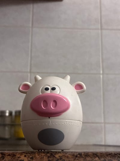 A close-up, eye-level shot shows a white cow-shaped kitchen timer sitting on a granite countertop. The timer is positioned centrally in the frame, facing forward. It has large, black eyes, a pink snout with two oval nostrils, and small pink inner ears. Two small, white horns protrude from the top of its head. The lower body of the timer displays time markings with numbers visible including "55", "0", and other unreadable digits. A dark grey oval shape is located on the lower part of the timer. In the blurred background, a tiled wall is visible with faint horizontal and vertical grout lines. To the left of the timer, on a shelf, are out-of-focus spice jars, one appearing to be clear and the others colored yellow and silver. The image appears to be taken indoors in what resembles a kitchen. The location in Гаранскі сельскі Савет, Belarus cannot be determined from the visual information provided in the image.