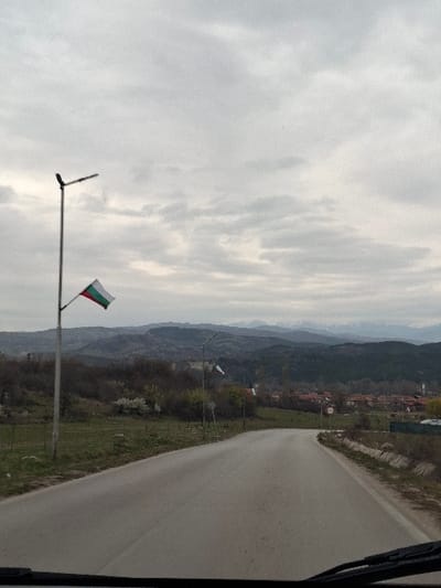 The image depicts a rural road scene in the city of Cherniche, Bulgaria, under an overcast sky. The perspective is from inside a vehicle, with the top of a dashboard and windshield wipers visible at the bottom edge. A paved asphalt road, showing signs of wear, curves slightly to the right, receding into the distance.

On the left side of the road, a tall, grey metal street lamp pole stands prominently, adorned with a Bulgarian flag featuring horizontal white, green, and red stripes, positioned beneath the luminaire arm. Further along the road on the left, another similar street lamp with a flag is visible, along with a verdant area comprising grass, shrubs, and scattered deciduous trees.

The right side of the road features a concrete or stone-lined drainage ditch. Beyond this, additional street lamps with Bulgarian flags are spaced along the roadside. In the midground, beyond the immediate roadside vegetation, a cluster of buildings with red-tiled roofs indicates a settlement.

The background is dominated by a series of undulating hills and mountains, some of which exhibit visible snow-capped peaks. The sky is entirely covered by grey, unbroken cloud cover, suggesting an overcast day. No individuals or active subjects are discernible in the frame.