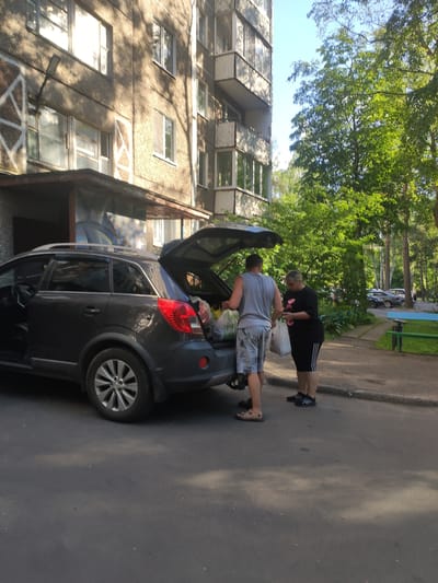 Two people are standing next to a dark-colored car with its trunk open. One person, wearing a black t-shirt with white stripes on the sleeves and black shorts, is reaching into the trunk. The other person, wearing a grey t-shirt and patterned shorts, is standing behind the car, also appearing to interact with the trunk. The car is parked on an asphalt surface, likely a street or parking lot.
Behind the car, there is a building with multiple windows, suggesting an urban or residential setting. Trees with green foliage are visible, indicating it is daytime and likely a warm season. The sky is bright, suggesting clear weather. The scene appears to be in Navapolatsk, Belarus. The overall impression is of people loading or unloading items from the car. No visible text is present.