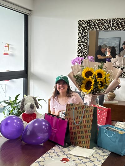 In Santa Clara Ocoyucan, Mexico, a young female, appearing to be in her late teens or early twenties, is seated at a dark wooden table, smiling directly at the camera. She is wearing a light pink t-shirt and a green baseball cap. To her immediate left on the table are two purple balloons and a white and brown plush dog, resembling Snoopy, adorned with a red heart bearing the text "SMILES." Directly in front of her are several gift bags: a pink bag partially displaying "Liverpool" branding, a large brown bag patterned with multiple green and red gift boxes, a red bag with white polka dots, and an open blue bag revealing contents within. Behind these gifts, a large bouquet of sunflowers wrapped in light brown paper, accented with baby's breath, is centrally placed, with a smaller bouquet of purple orchids visible to its left. A white and blue patterned table runner, a white plate with a red poppy design, and a folded white napkin are also present on the table. The room features white walls. A large window with a black frame and a white roller blind is positioned on the left, through which a green plant and a red-accented hummingbird feeder are visible. On the right wall, a large mirror with an ornate, geometric brown and white frame reflects a grey-haired man in a black shirt embracing a dark-haired woman, with additional individuals partially visible in the reflection. A dark wooden credenza, holding a vase and books, is situated beneath the mirror. The scene depicts an indoor celebratory gathering.